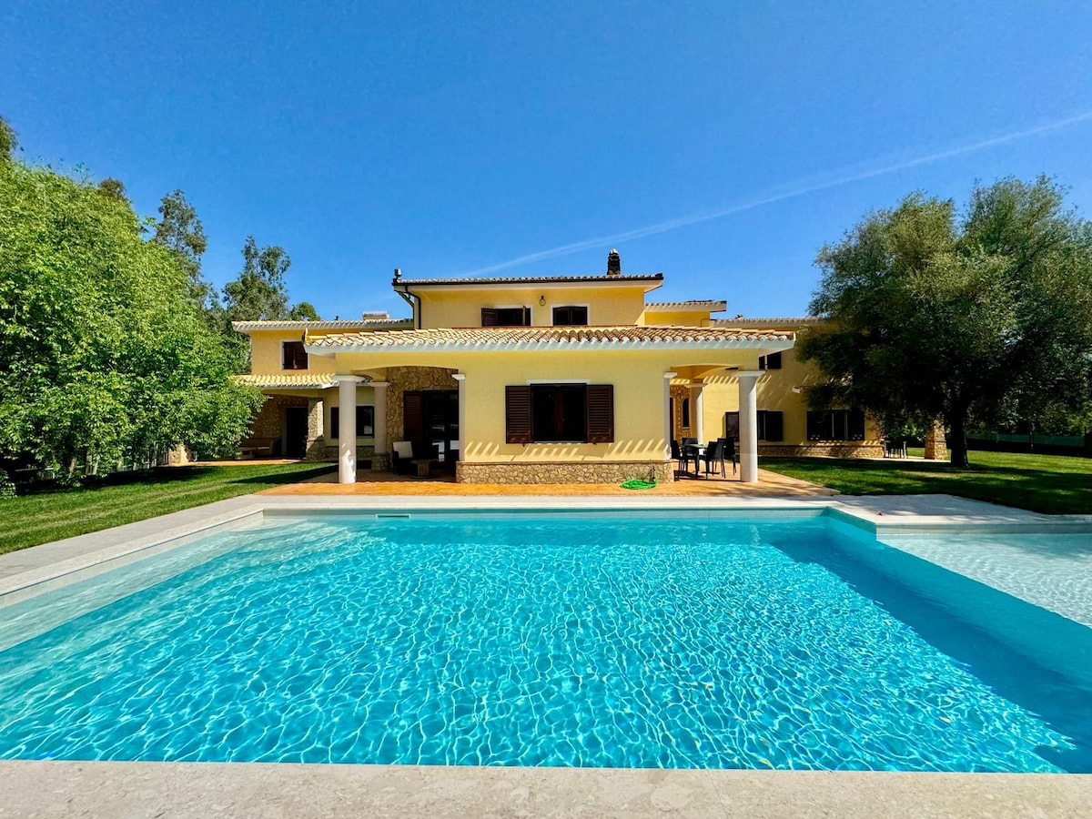 The villa features a spacious outdoor area with a shared swimming pool surrounded by well-maintained green grass and trees. The building showcases a two-story structure with large windows and a terrace area for relaxation. Clear skies provide a bright backdrop against the yellow façade.
