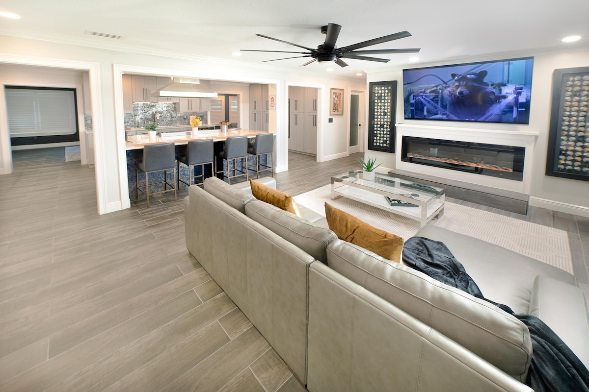 An open living area features a spacious sectional sofa facing a large flat-screen TV mounted above a modern fireplace. The kitchen area, visible in the background, includes a sleek countertop with bar seating. Natural light highlights the room, enhancing the contemporary design.