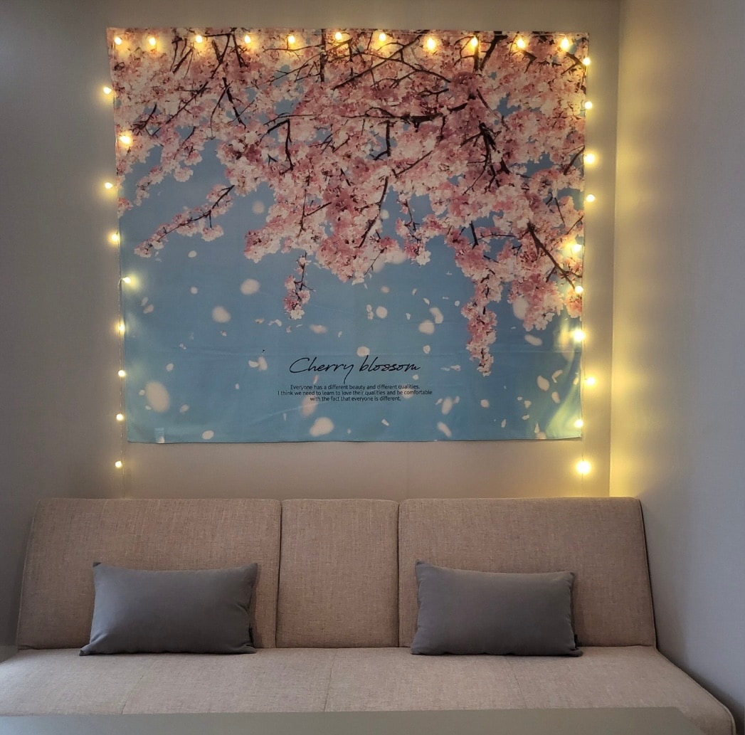 A cozy seating area is highlighted by a large wall tapestry featuring cherry blossoms against a sky-blue background. Soft lighting strands are draped around the tapestry, creating a serene ambiance. Two cushions in muted colors are placed on a light-colored sofa.
