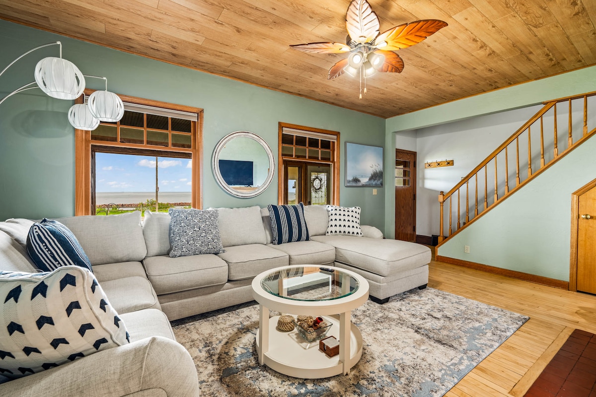 A spacious living area is highlighted by a large sectional sofa adorned with blue and white accent pillows. A circular coffee table sits on a soft area rug. Natural light fills the room through expansive windows, offering views of the outdoors. Light wood paneling is featured on the ceiling.