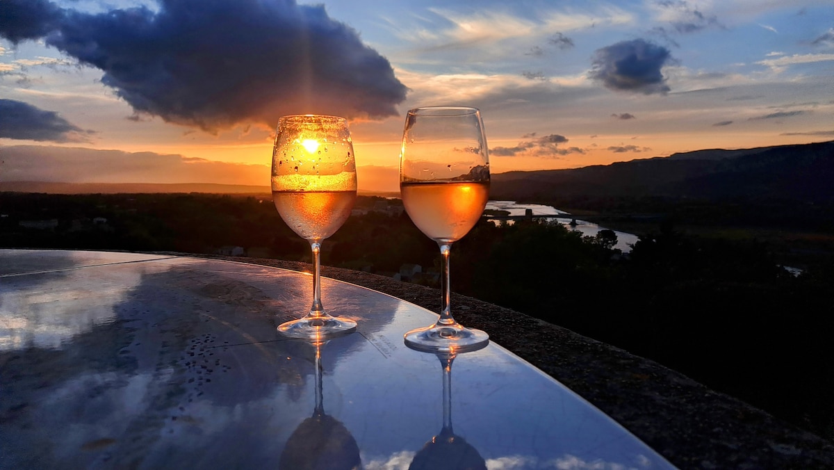 Two glasses of wine are placed on a reflective surface, capturing the colors of a sunset. The sky transitions from warm orange and pink tones to cooler blues, with distant hills and a river in the background, creating a serene and picturesque view.