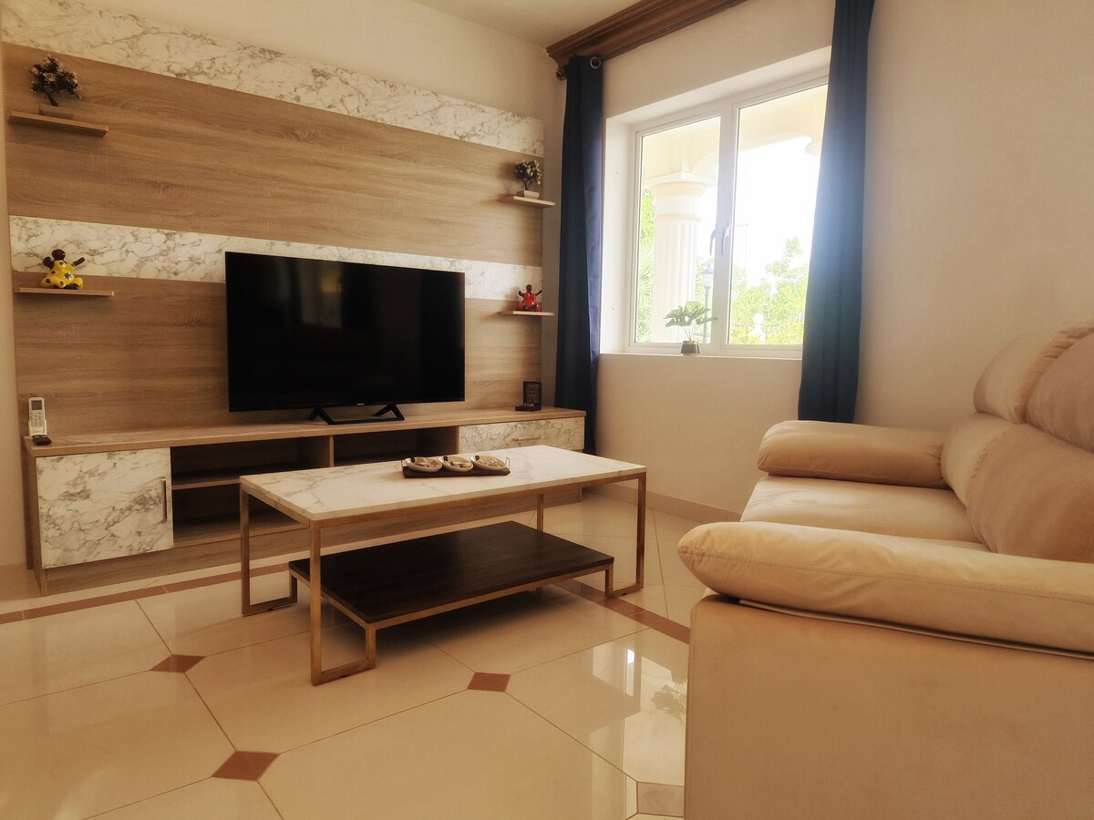 A light-filled living area is presented, featuring a mounted flat-screen TV on a marble-accented wall. A stylish coffee table rests on the glossy tiled floor, complemented by a cream-colored sofa. Natural light enters through a window adorned with simple curtains.