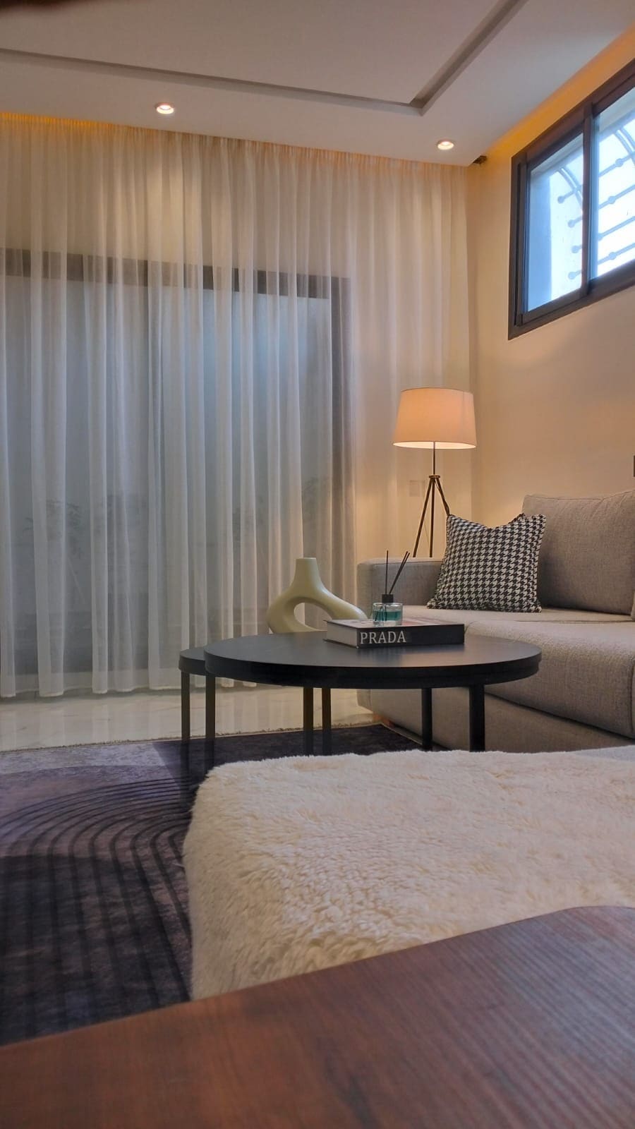 A comfortable living area is presented, featuring a plush sofa and a round coffee table. Soft, sheer curtains frame a window, allowing natural light to filter through. A stylish lamp stands beside the sofa, enhancing the space's modern aesthetic.