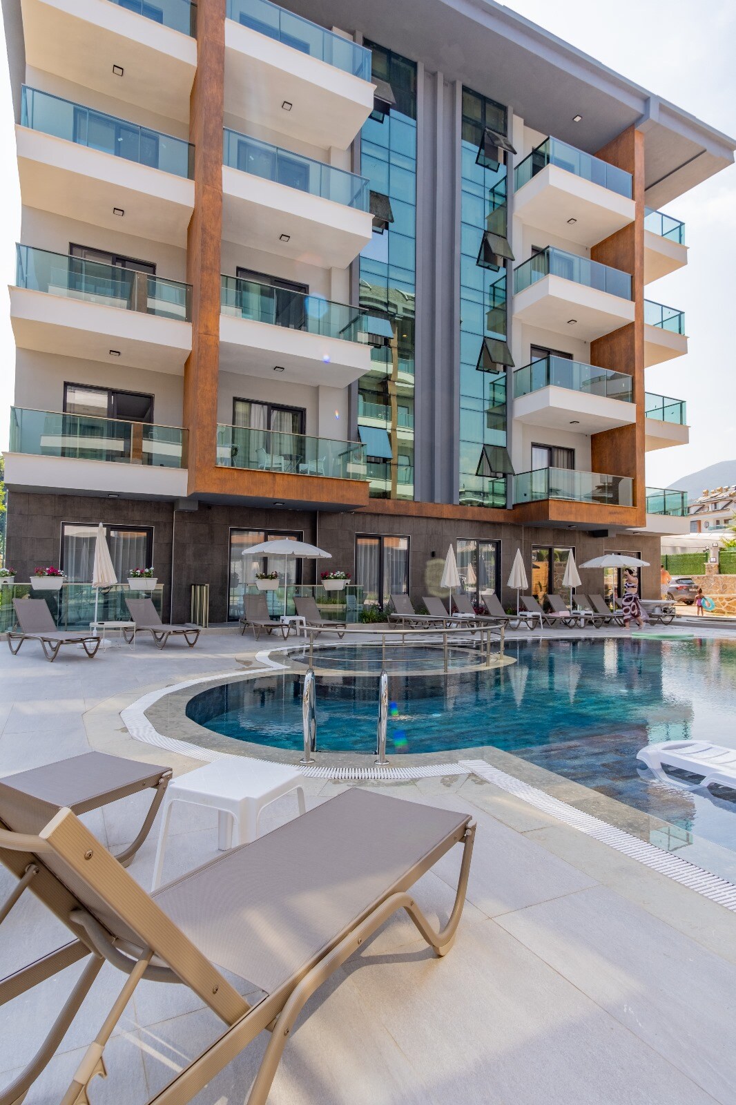 The modern building features multiple floors with glass balconies. A tranquil pool is surrounded by lounge chairs and umbrellas, creating a welcoming outdoor space. The structure's contemporary design is complemented by wood accents and large windows reflecting the surrounding area.