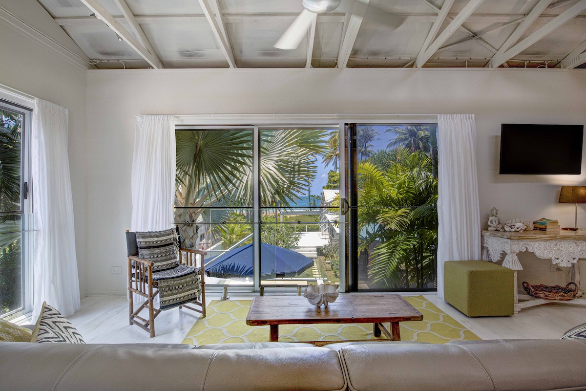 Natural light fills the spacious living area through large glass windows, providing views of swaying palm trees and the ocean. A comfortable couch faces a wooden coffee table, while a stylish chair adds seating. Decorative elements on the side table complement the serene atmosphere.