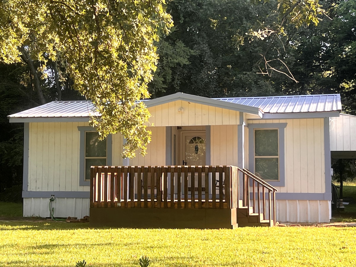 The exterior of the lake house is shown, featuring a welcoming front porch with a brown railing. A light-colored facade is complemented by gray trim. Lush green grass surrounds the home, with trees providing shade in the background.