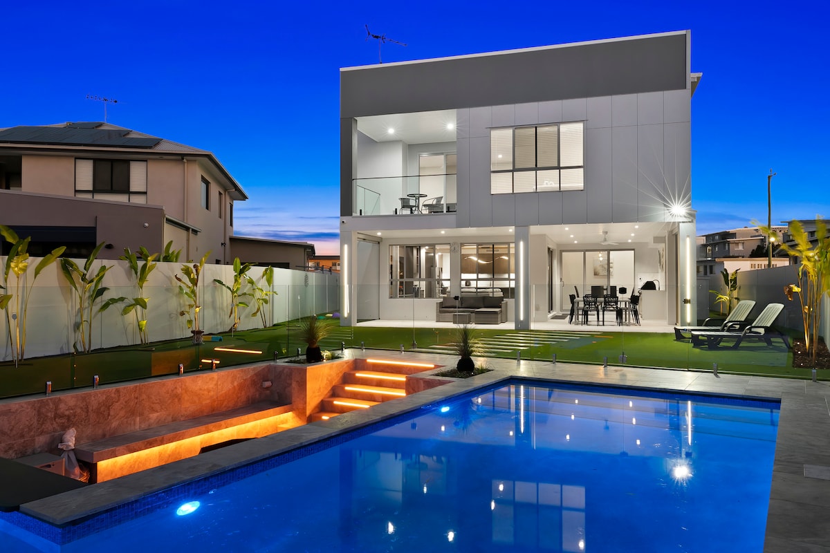A modern holiday home is displayed against a twilight sky, featuring a crystal blue inground pool. The exterior showcases a spacious open-plan layout with large glass doors leading to the outdoor dining area. A hot tub is situated beside the pool, with subtle lighting enhancing the scene.