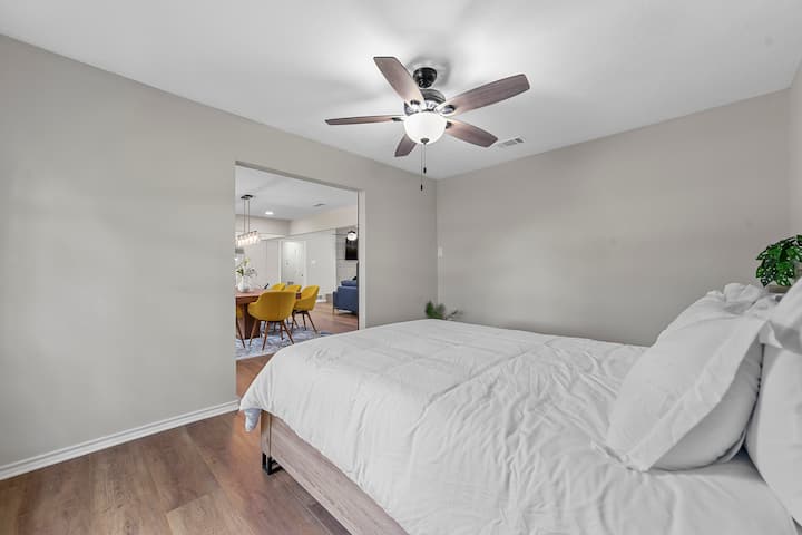 Dhomë gjumi e ndritshme me një krevat të bardhë, dysheme druri dhe ventilator modern tavani. Dritaret e mëdha me hark lejojnë hyrjen e dritës natyrale. Dhoma hapet në një hapësirë elegante ngrënieje me karrige të verdha, duke përzier rehatinë dhe dizajnin në një planimetri të përsosur.