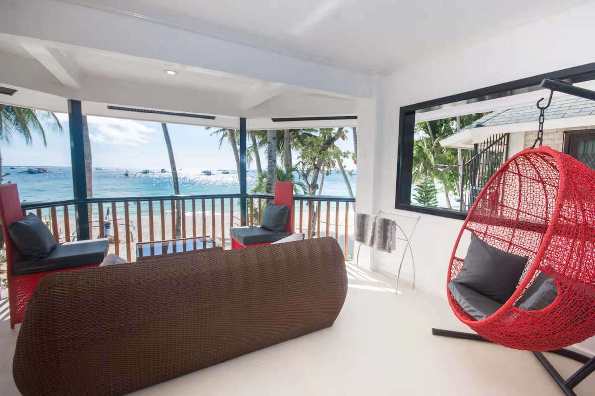A brightly lit seating area offers a view of the beach through large windows. Stylish chairs and a unique hanging chair are arranged around a modern table. Lush palm trees can be seen in the background, enhancing the coastal ambiance.