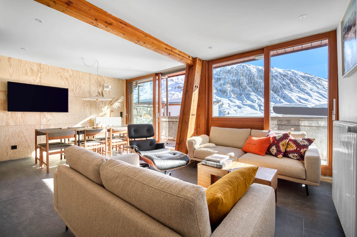 A spacious living area showcases large windows framing stunning mountain views. Contemporary seating arrangements include a comfortable sofa and armchair, complemented by a wooden coffee table. A sleek television is mounted on the wall, enhancing the inviting atmosphere of the space.