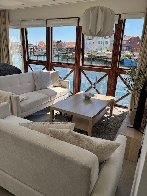 A light-filled living area features two comfortable sofas facing a wooden coffee table. Large windows provide an unobstructed view of the waterfront, allowing natural light to illuminate the space. Neutral tones are complemented by decorative accents, creating a cozy environment.