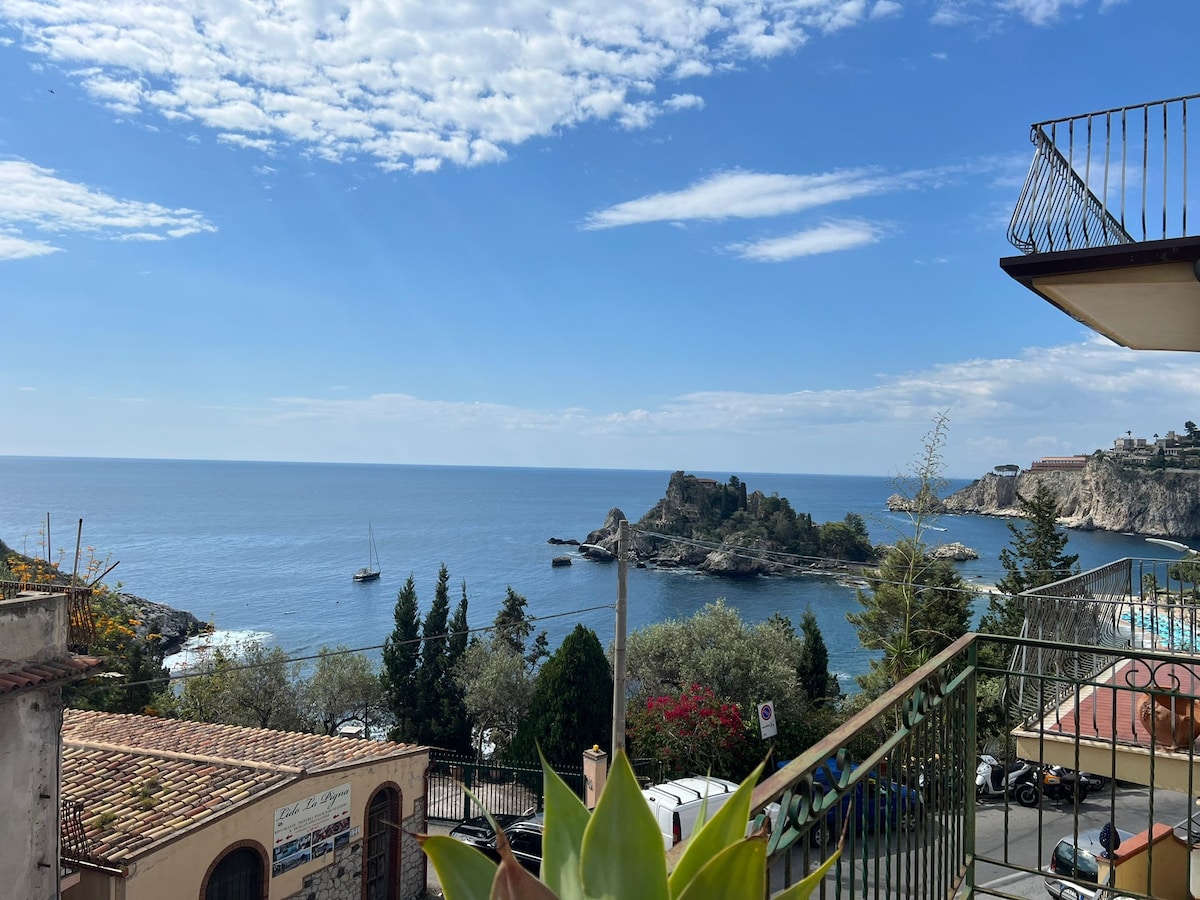 A tranquil view of the waterfront is captured from the terrace, showcasing serene blue waters and lush greenery. A historic structure is visible on the rocky outcrop, surrounded by a calm sea. The sky is bright with scattered clouds, enhancing the coastal atmosphere.