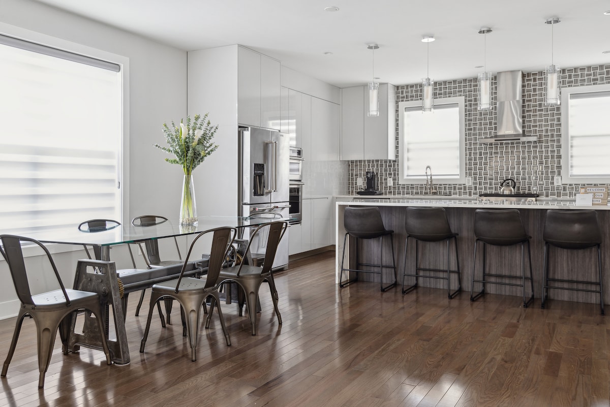 A modern kitchen and dining area features sleek cabinetry and stainless steel appliances. A glass dining table is surrounded by black metal chairs. The bar counter, fitted with high stools, complements the hardwood flooring and mosaic tile backsplash.