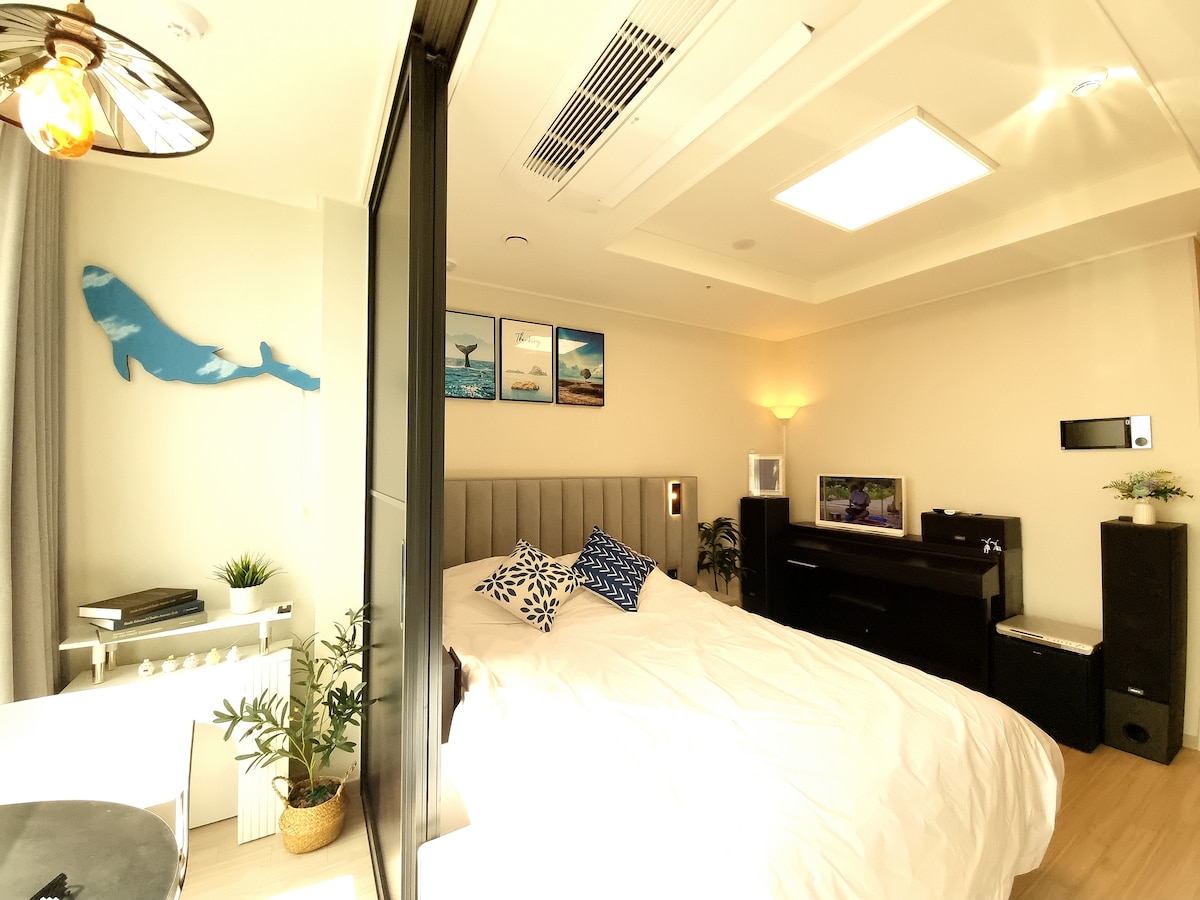 A spacious sleeping area features a large bed adorned with decorative pillows. Decorative artwork depicting marine life is displayed on the wall. A soothing glow is provided by various lighting elements, while a modern entertainment unit and potted plants add to the room's ambiance.