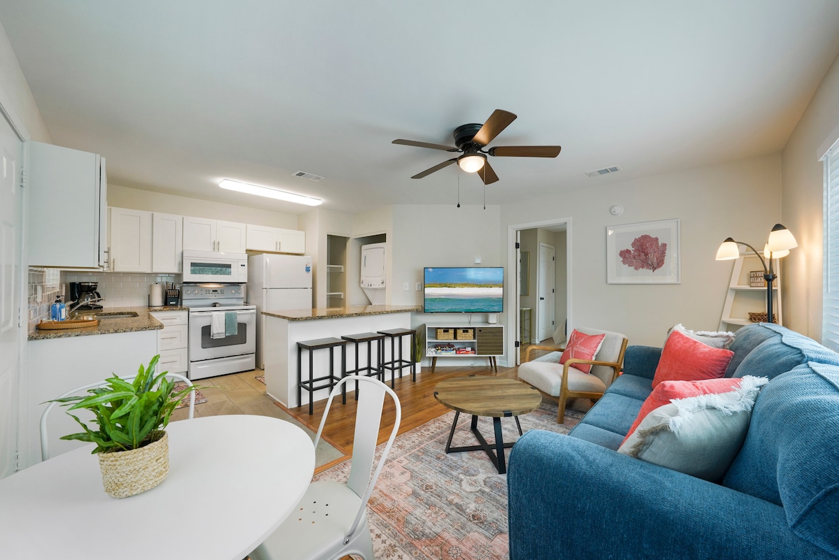 A bright and airy living space showcases an open layout with a comfortable blue sofa adorned with colorful pillows. A round dining table with white chairs complements the modern kitchen, which features white cabinetry and stainless steel appliances, enhancing the inviting atmosphere.