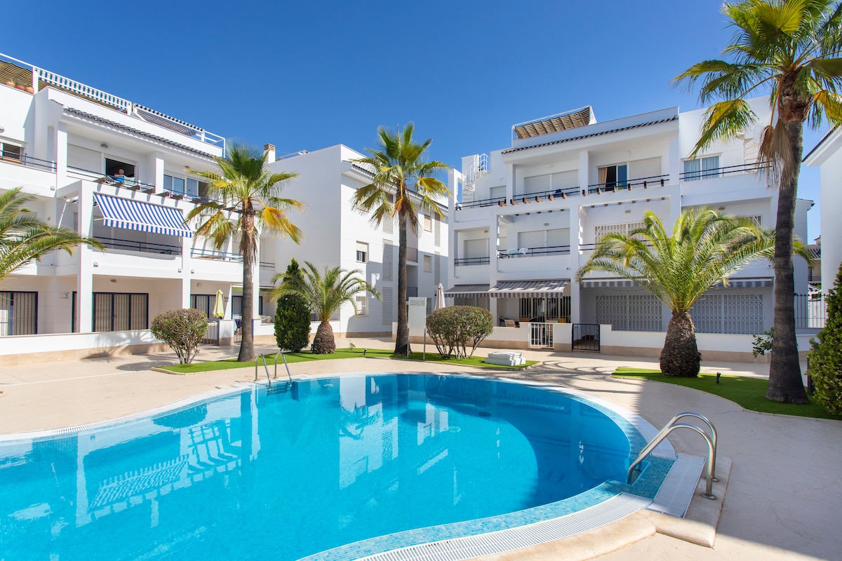 A well-maintained common area features a clear blue pool surrounded by lush palm trees. The modern buildings stand in the background, with balconies visible. The tranquil setting suggests a relaxed atmosphere, ideal for enjoying sunny days by the water.