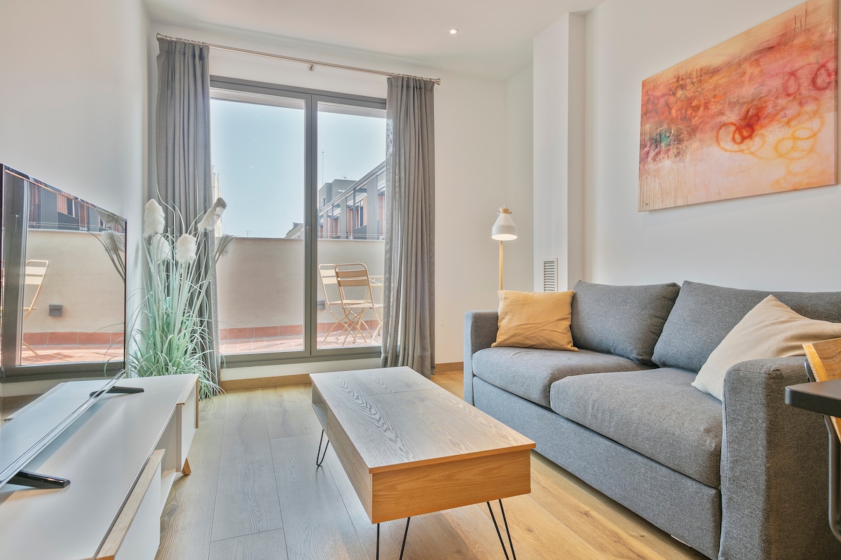 A light-filled living area features a gray sofa adorned with a soft pillow, positioned near a coffee table made of wood. Large glass doors open to a balcony, revealing outdoor seating. A TV is mounted on a stand, completing the inviting arrangement.