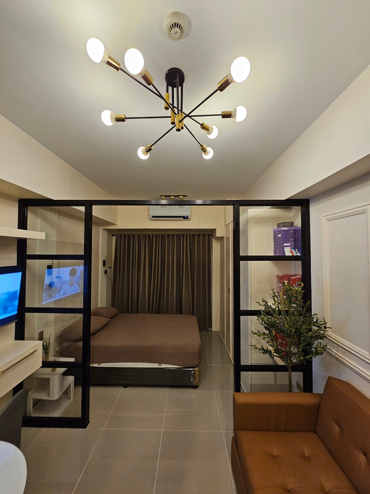 The living space features a minimalist design with a spacious bed and a comfortable sofa. A modern chandelier hangs from the ceiling, while glass partitions separate the sleeping area from the living area. Light brown curtains frame a window, enhancing the cozy atmosphere.