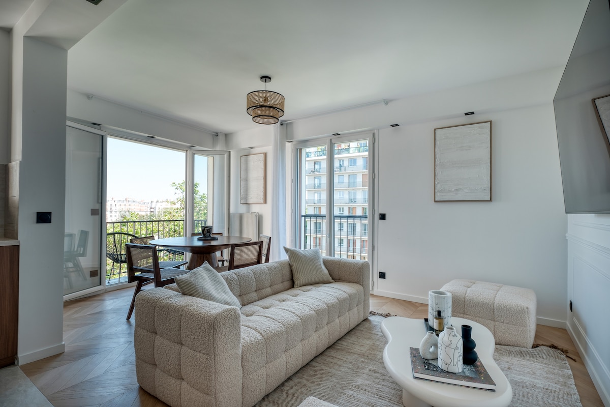 A light-filled living area features a spacious beige sofa and a minimalist coffee table, set against a backdrop of large glass doors. A dining table with four chairs provides an inviting space for meals, while a neutral palette enhances the room's modern design.