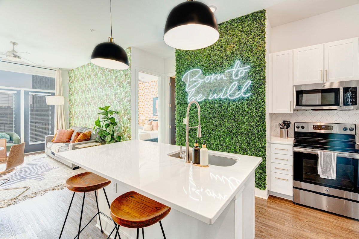 An inviting kitchen features a sleek island equipped with two wooden bar stools. A neon sign reading 'Born to be Wild' adds a modern touch, complemented by a backdrop of vibrant greenery. Stainless steel appliances and white cabinetry contribute to a clean and contemporary aesthetic.