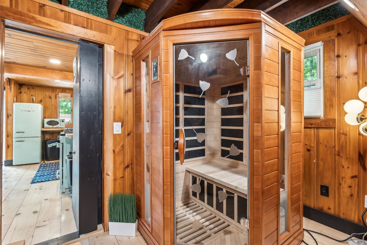 An infrared sauna is presented within a wooden interior, featuring a glass-enclosed design. The sauna is equipped with wooden benches and heart-shaped detailing on the interior walls. Natural light filters in through nearby windows, enhancing the relaxing atmosphere of the space.