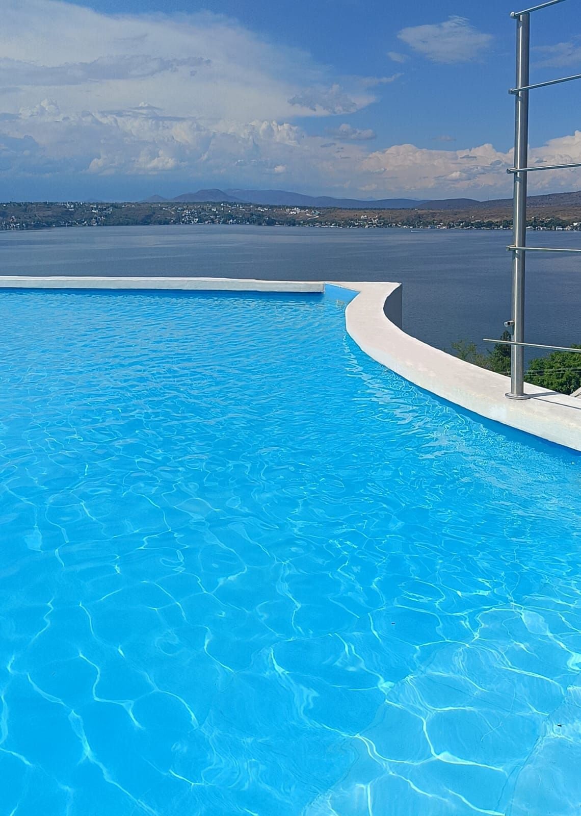 An infinity pool extends towards the horizon, featuring clear, shimmering water that reflects the sky. The landscape in the background includes a lake bordered by rolling hills and distant mountains, adding a sense of openness to the serene setting.