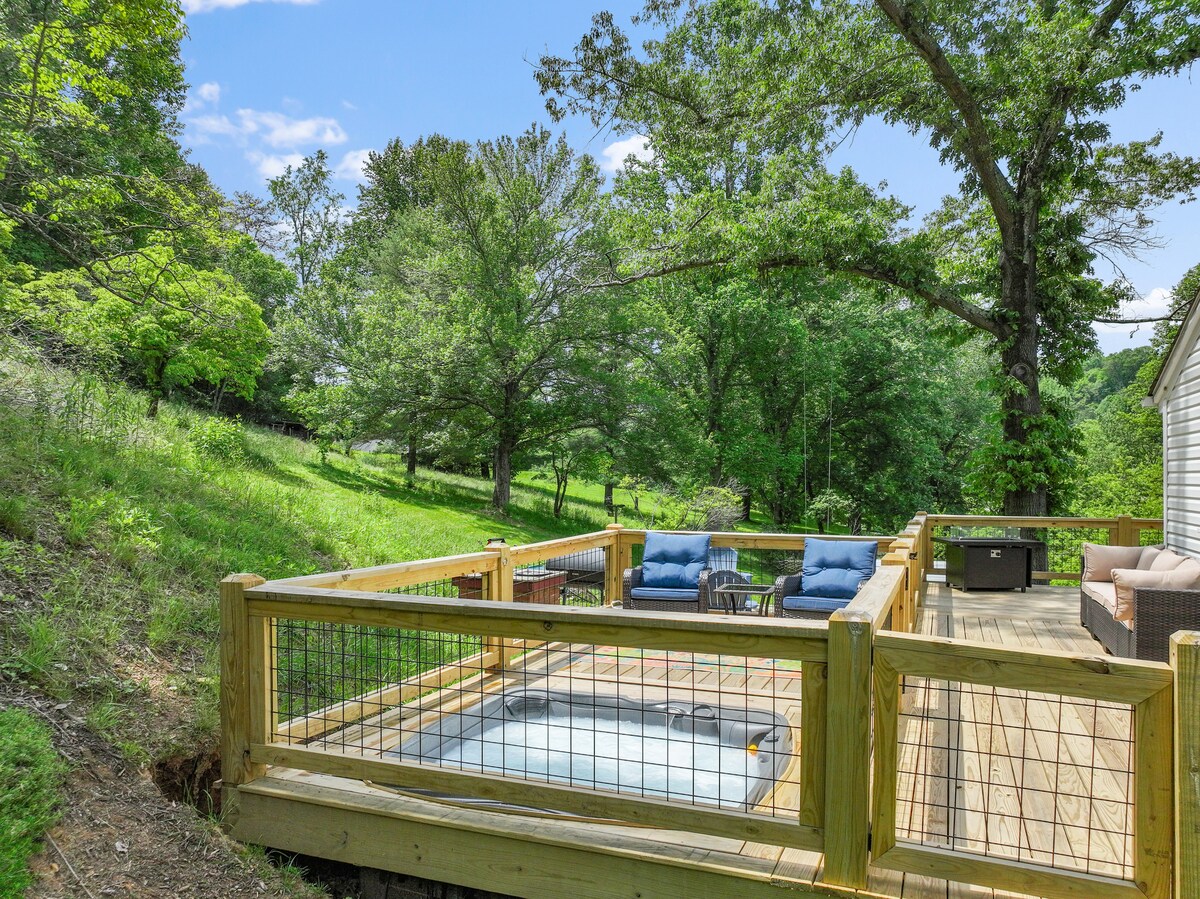 A wooden deck features a hot tub surrounded by protective railing. Two cushioned chairs and a small table are positioned nearby, providing space to relax. Lush greenery and trees extend into the background, enhancing the natural setting of the property.