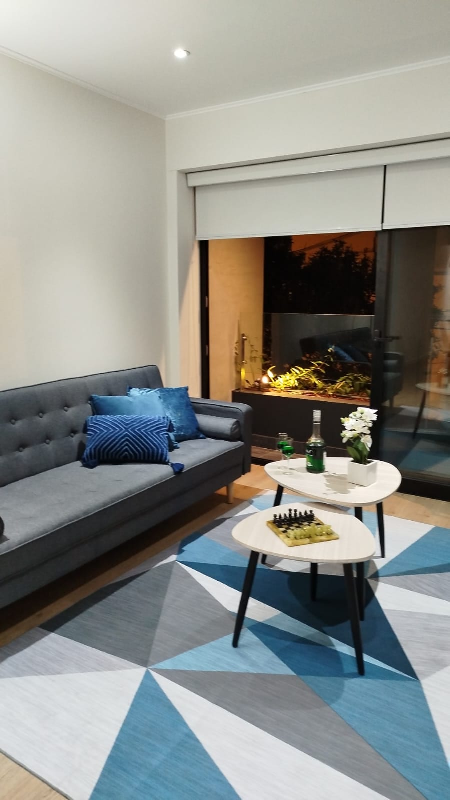 A modern living area features a gray sofa with decorative blue pillows, positioned near a glass balcony door. A round coffee table sits in front, displaying a bottle of champagne, glasses, and a small floral arrangement. A geometric area rug adds a touch of color.
