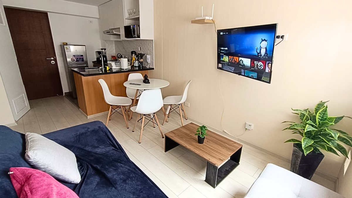 A modern living space features a comfortable sofa adorned with colorful cushions. A small dining table with four chairs is positioned near an open kitchen. A wall-mounted TV displays a screen, and indoor plants add a touch of greenery to the area.