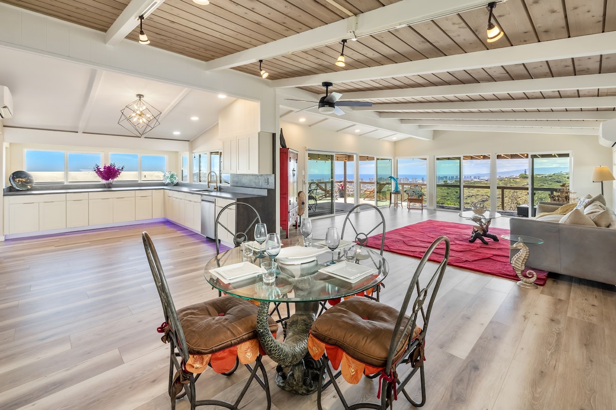 An open-concept living area features a spacious kitchen and dining space. A glass-top dining table surrounded by metal chairs is set for a meal. Large windows offer panoramic views of the surrounding landscape, while a comfortable seating area includes a neutral-toned sofa and colorful area rug.
