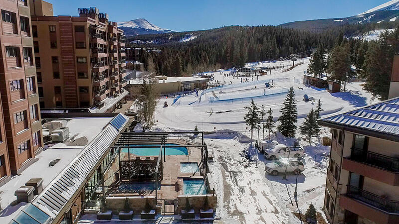 A panoramic view showcases the snow-covered landscape, with ski slopes and a winter sports area in the background. The image includes nearby buildings with a contemporary design. Pool areas and outdoor spaces are visible, framed by evergreen trees and a clear blue sky.