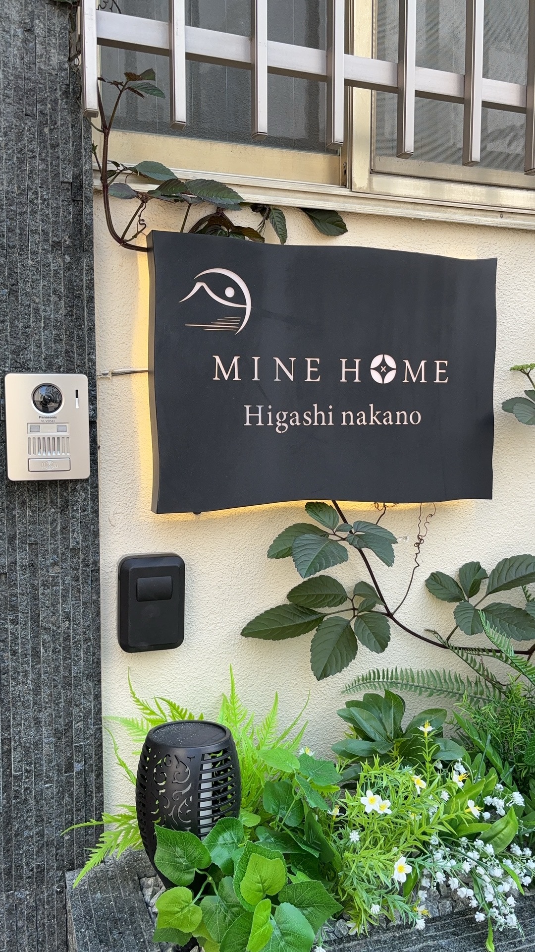 The entrance of the property is marked by a welcoming sign displaying 'MINE HOME Higashi Nakano.' Lush green plants and decorative accents are arranged around the sign, contributing to a pleasant and inviting atmosphere.