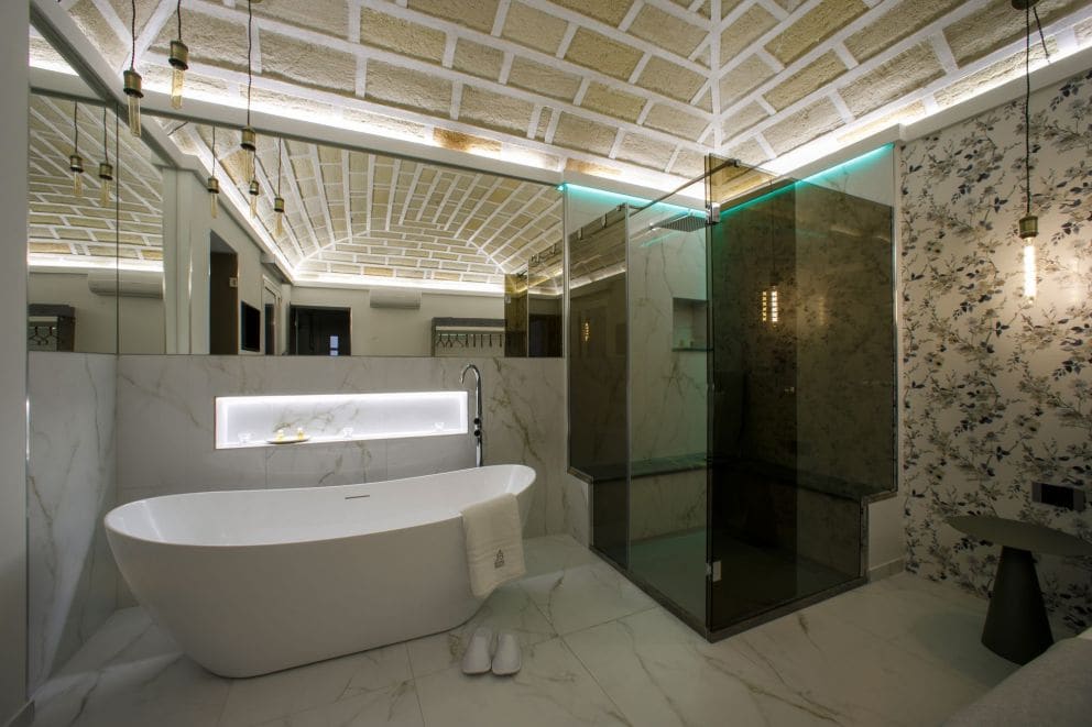 A modern bathroom showcases a freestanding bathtub accompanied by plush towels. The sleek glass shower is positioned beside textured marble walls, while stylish pendant lights hang from the ceiling along a patterned design. Soft lighting enhances the space's tranquil atmosphere.