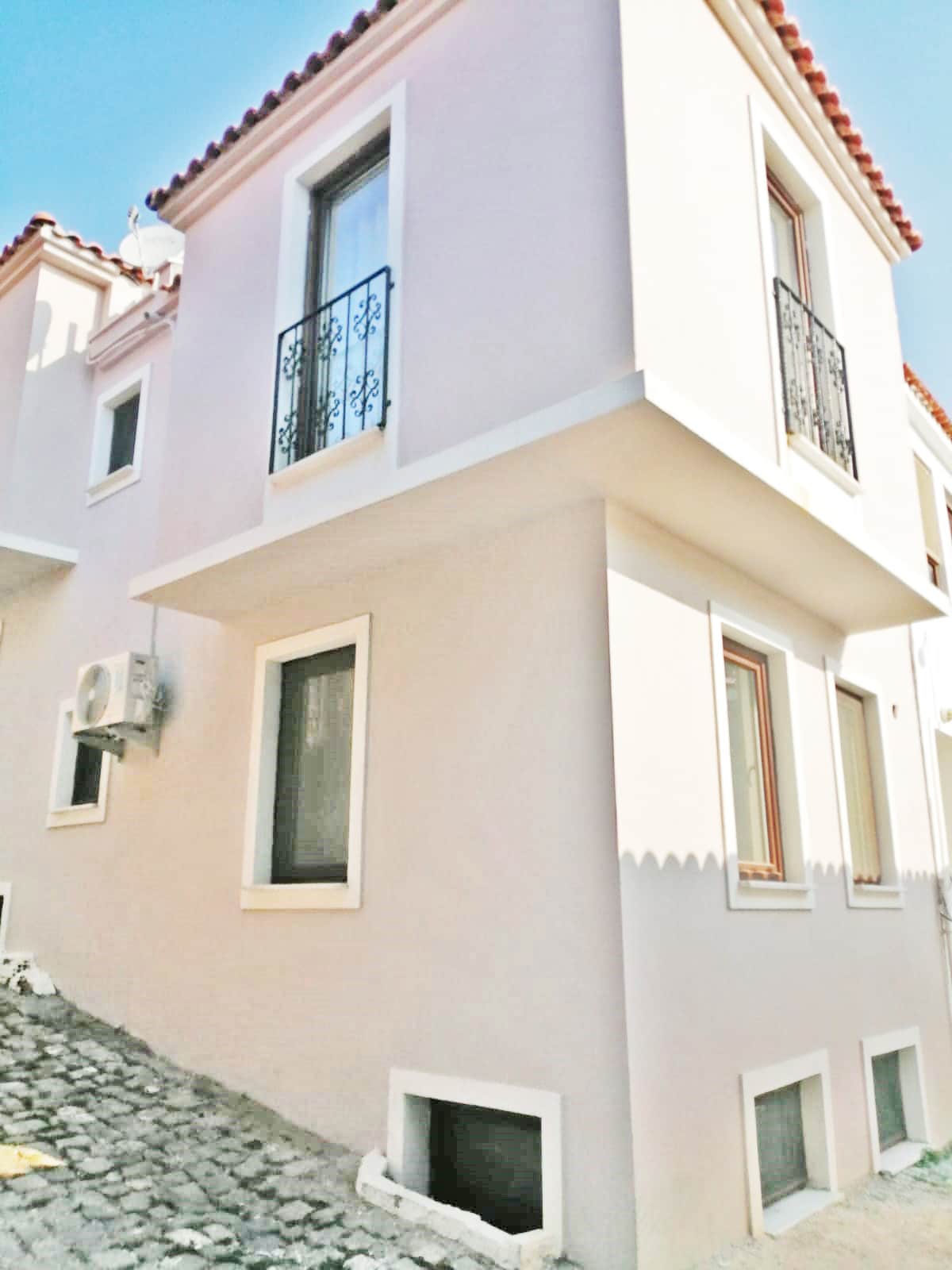 The exterior of a two-story building features a clean stucco façade complemented by decorative wrought-iron balconies. Large windows provide natural light, and an air conditioning unit is mounted on the side. The surrounding cobblestone pathway enhances the building's charming appearance.