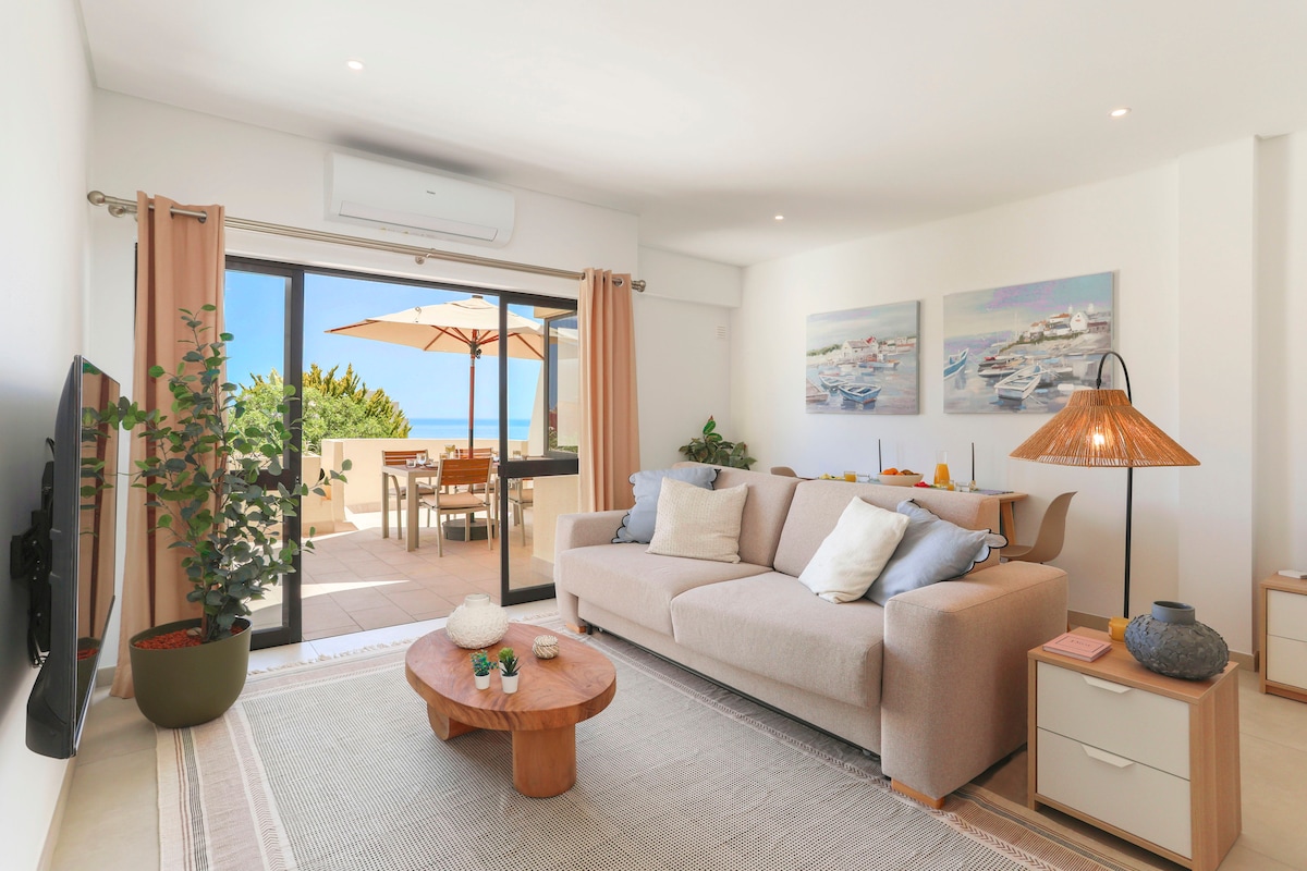 A spacious living room features a light-colored sofa, a round wooden coffee table, and large windows that open to a balcony with ocean views. The room is enhanced by light curtains and decorative wall art, creating a bright and welcoming ambiance.
