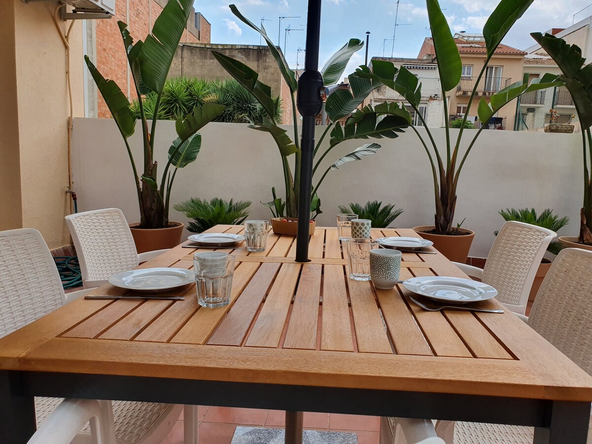 A spacious terrace features a wooden dining table with matching chairs, set for a meal. Glassware and tableware are neatly arranged on the table. Surrounding greenery, including large potted plants, enhances the outdoor setting, providing a calming atmosphere.