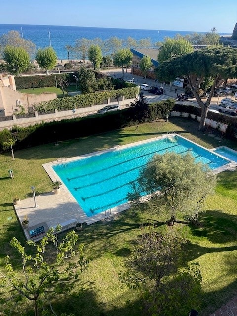 A clear view of a private swimming pool surrounded by well-maintained gardens. The water appears inviting under sunlight, and tall trees offer shade nearby. In the background, the sea is visible, enhancing the serene environment.