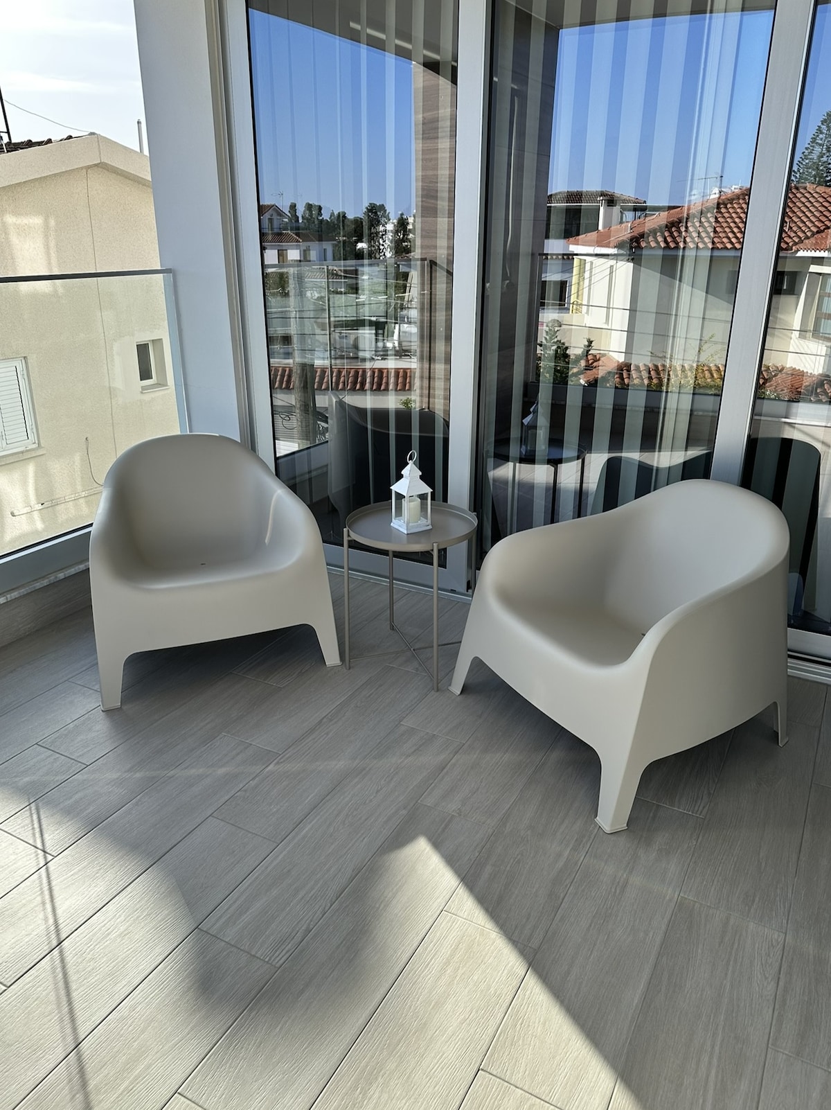 A corner of the balcony is furnished with two modern chairs and a small round table. Natural light fills the space, highlighting the light-colored tiled flooring and the surrounding views through large glass sliding doors.