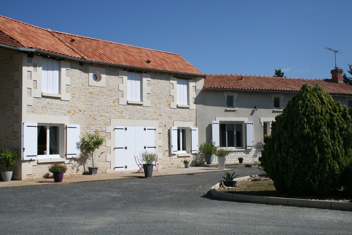 A charming stone property is visible, featuring white shutters, a red-tiled roof, and a pebbled driveway. Surrounded by neatly trimmed hedges and potted plants, the peaceful exterior conveys a sense of tranquility. Clear blue skies provide a bright backdrop to the inviting entrance.
