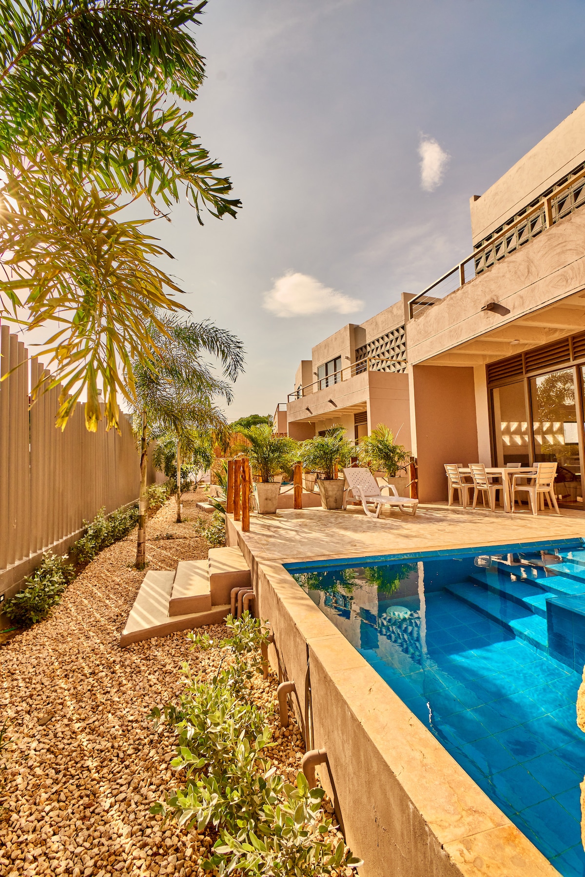 A spacious outdoor area features a private swimming pool surrounded by lush greenery and decorative stones. Sunlight filters through palm trees, illuminating the terrace area with lounge chairs and a dining set. The modern cabañas are visible in the background, providing a relaxed atmosphere.