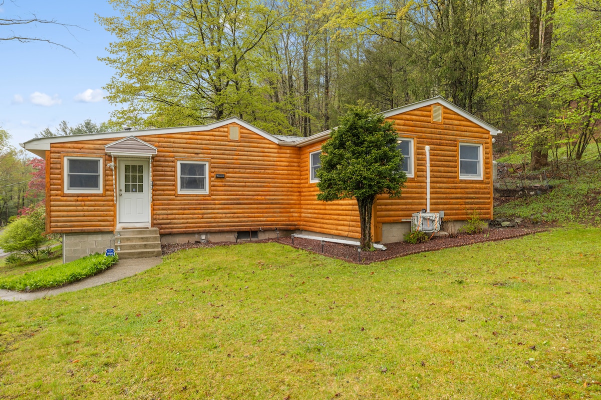 A cozy cabin exterior is displayed, featuring warm wooden siding that blends with the surrounding greenery. The well-maintained lawn offers a peaceful setting, while a walkway leads up to the front door framed by a small tree and shrubs.