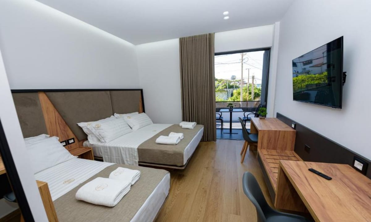A stylish guest room features two spacious beds with neutral-toned bedding and wooden headboards. A sleek desk and a chair are positioned against one wall. Large windows allow natural light to enter, with a view of a small balcony visible outside.