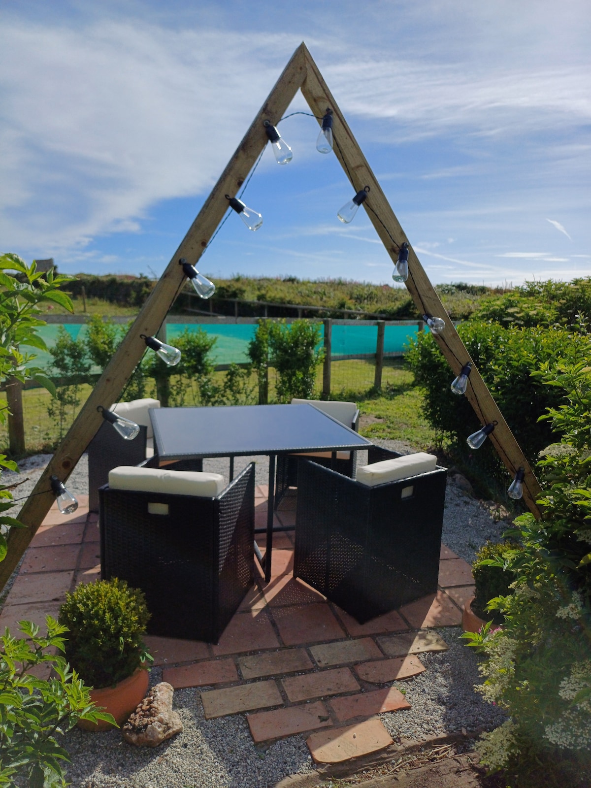 A triangular wooden structure adorned with string lights provides a cozy dining space. Two chairs and a table are situated on a terracotta patio surrounded by greenery. A scenic view of the landscape is visible in the background under a clear blue sky.