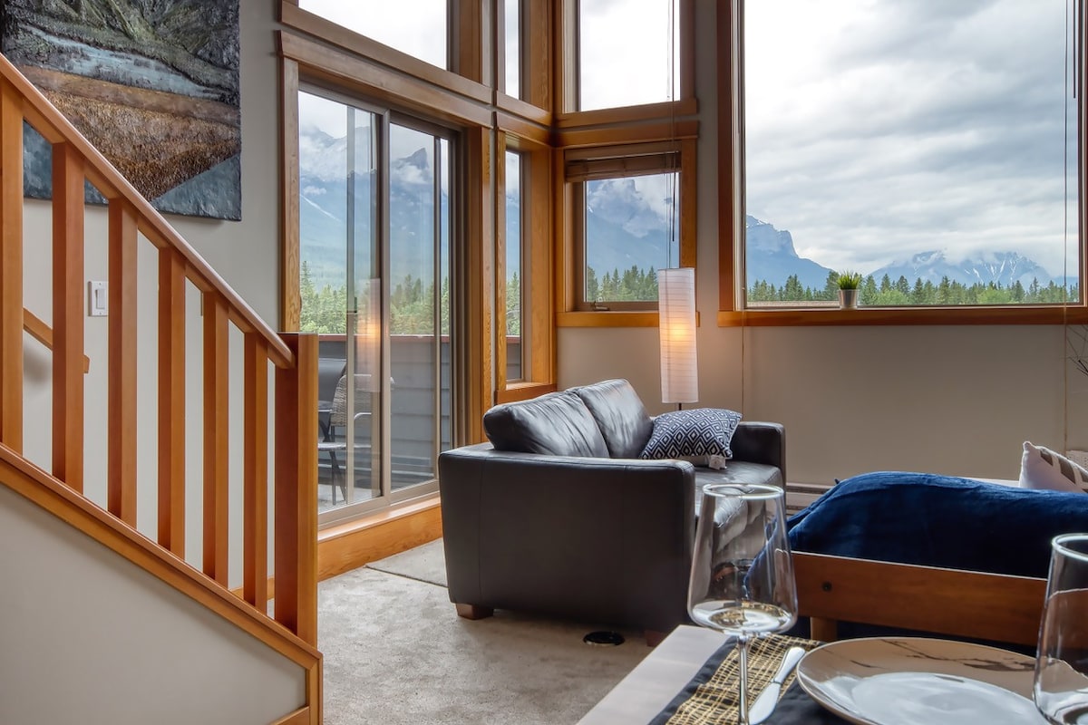 The living area features a comfortable leather sofa positioned near large corner windows, showcasing panoramic mountain views. A wooden staircase leads to the upper level. A glass dining table is set for two, with elegant tableware visible in the foreground.