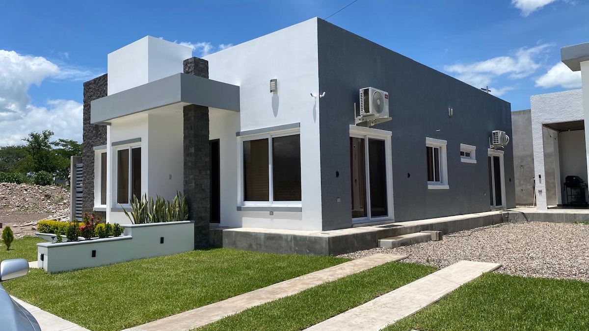 A modern single-story home features a combination of light and dark exterior walls. Landscaped grounds with green grass and flower beds are visible. The structure includes multiple windows and air conditioning units, offering a spacious and well-lit entrance.
