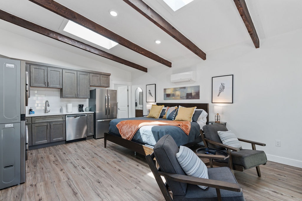 The studio features tall ceilings with exposed wooden beams and two skylights providing abundant natural light. A spacious king bed is positioned in the center, complemented by a kitchenette, stainless steel appliances, and two leather chairs for seating. Modern art decorates the walls.