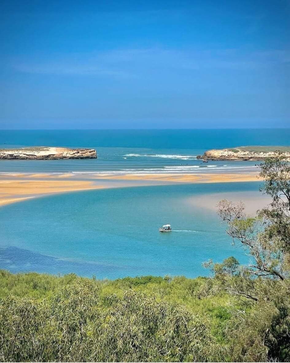 A panoramic view of the coastline is presented, featuring calm blue waters blending with sandy shores. Gentle waves lap against the beach, and a small boat glides through the tranquil bay. Lush greenery frames the scene, enhancing the natural beauty of the seascape.