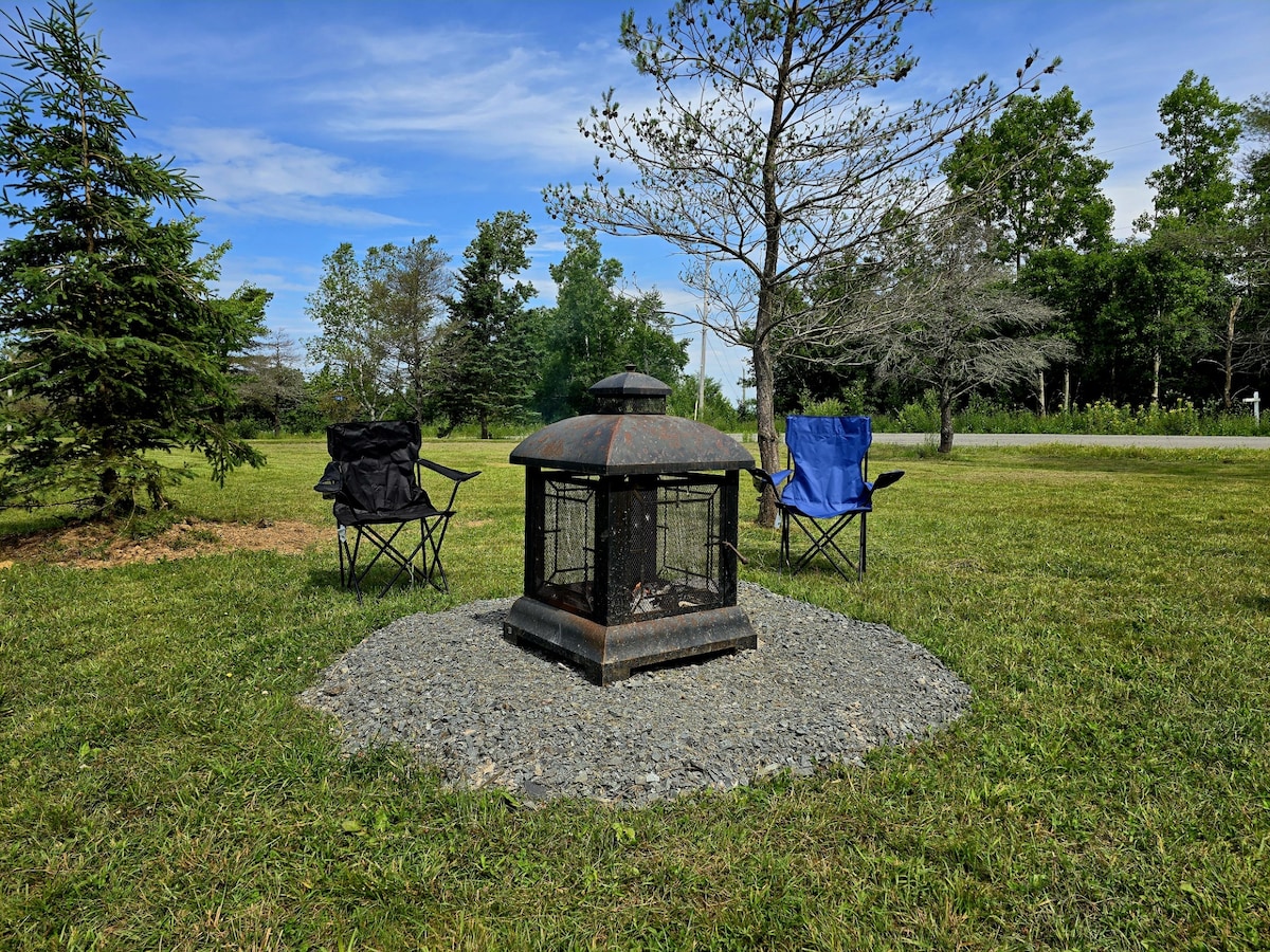 A fire pit is situated at the center of a stone circle in an open grassy area. Two folding chairs, one blue and one black, are positioned on either side of the fire pit. Surrounding trees provide a natural backdrop.