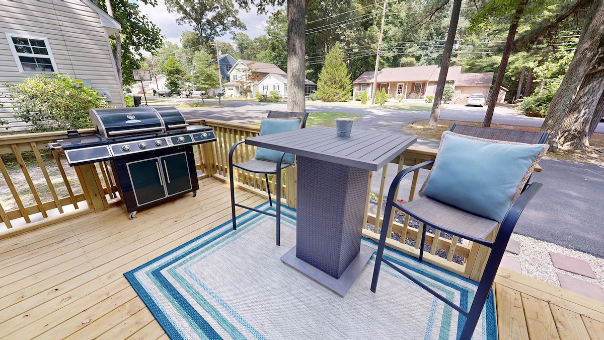 A wooden deck is presented with a small table and two seating chairs, complemented by a rug underneath. A grill is set nearby, while surrounding trees and houses create a serene neighborhood backdrop. The setup is ideal for enjoying outdoor meals.