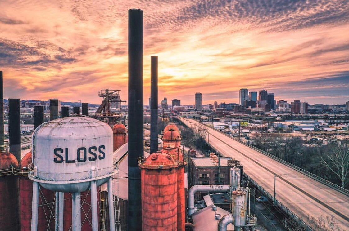 An industrial landscape captures a sunset view behind an old water tower labeled 'SLOSS', with smokestacks rising prominently. The city skyline is visible in the background, complemented by a highway stretching along the scene. Soft clouds color the sky in shades of orange and purple.